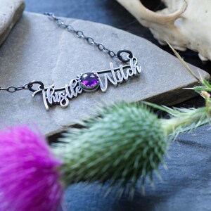 May include: A silver necklace with a purple gemstone and the words "Thistle Witch" written in cursive.