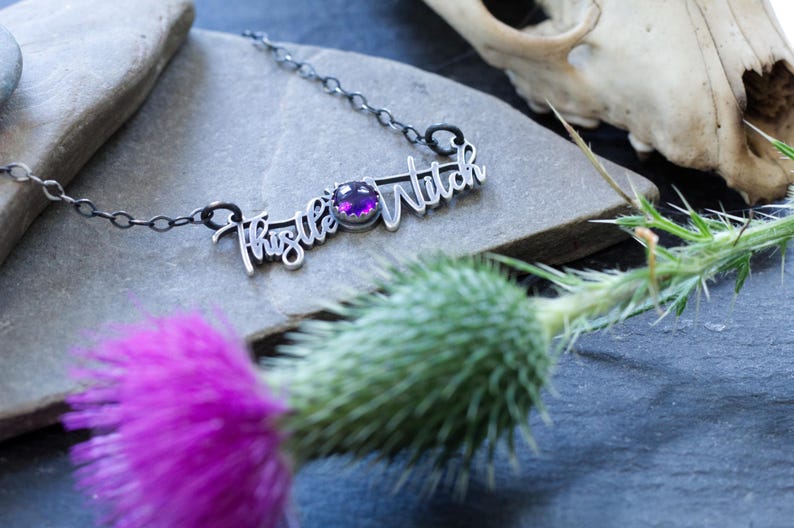 May include: A silver necklace with a purple gemstone and the words "Thistle Witch" written in cursive.