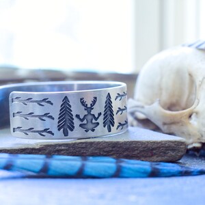 May include: A silver bracelet with an engraved design of trees, branches, and a figure with antlers. The bracelet is on a gray surface with a blue feather.