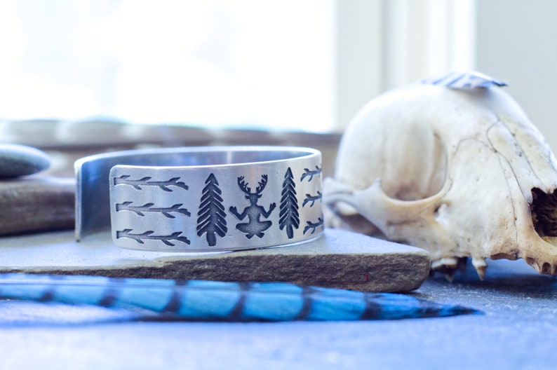 May include: A silver bracelet with an engraved design of trees, branches, and a figure with antlers. The bracelet is on a gray surface with a blue feather.