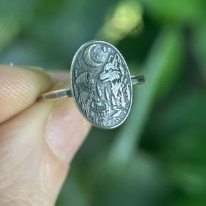 May include: A silver ring with an oval-shaped face featuring a detailed relief of a wolf howling at the moon. The ring has a thin band and a nature-inspired theme. The background is blurred green.