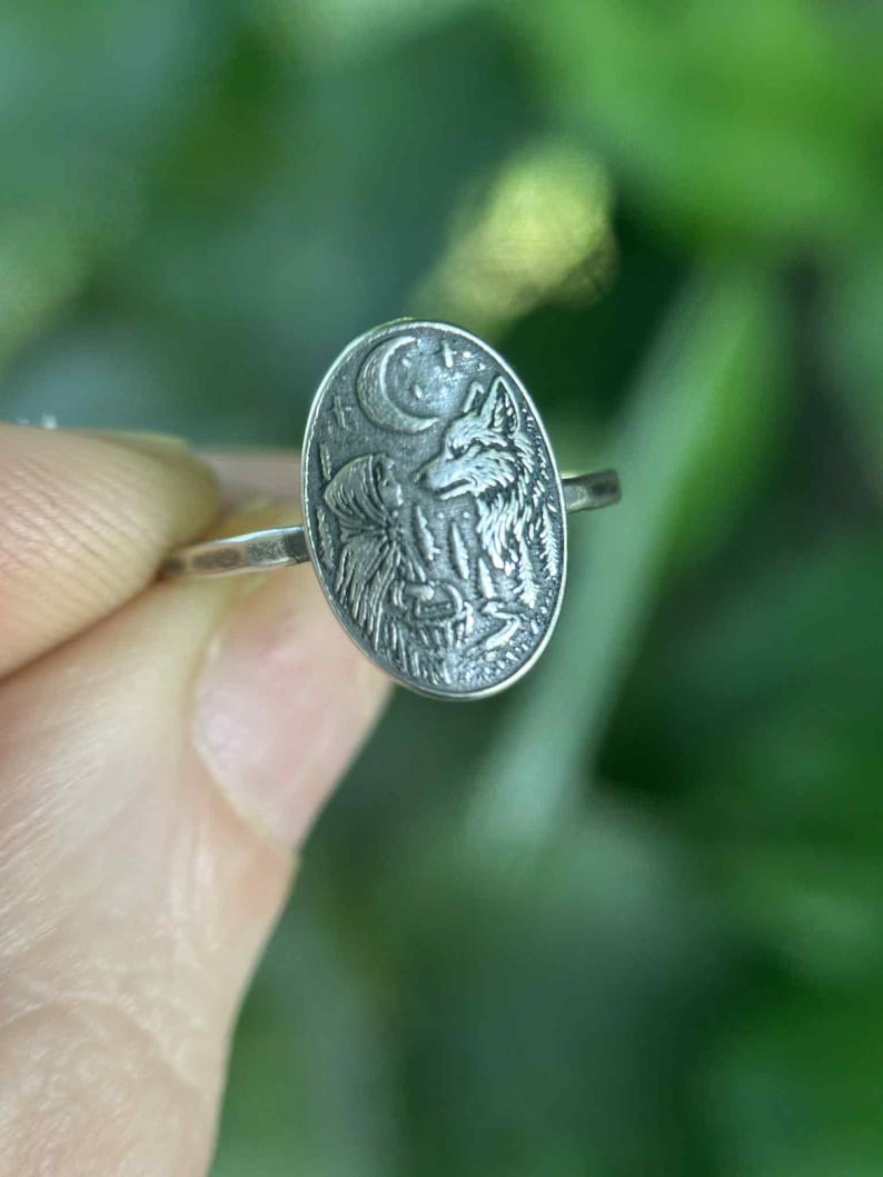May include: A silver ring with an oval-shaped face featuring a detailed relief of a wolf howling at the moon. The ring has a thin band and a nature-inspired theme. The background is blurred green.