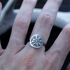 May include: A silver ring with a circular design featuring a compass rose with runes. The ring is worn on a person's finger.