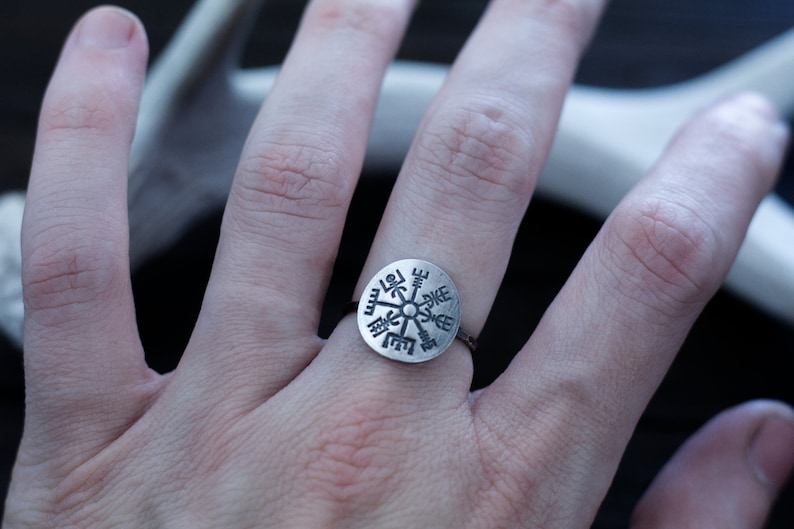 May include: A silver ring with a circular design featuring a compass rose with runes. The ring is worn on a person's finger.
