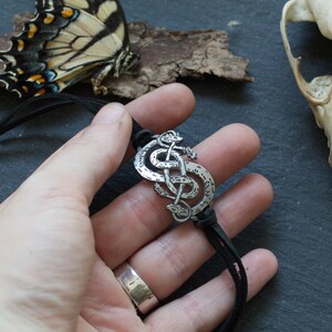 May include: A silver pendant necklace with a Celtic knot design featuring two intertwined snakes. The necklace is on a black leather cord.