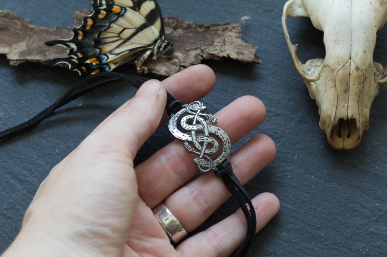 May include: A silver pendant necklace with a Celtic knot design featuring two intertwined snakes. The necklace is on a black leather cord.