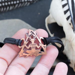 May include: A copper-colored bracelet with a leather cord and a pendant featuring a stylized figure with antlers, sitting in a triangular shape.