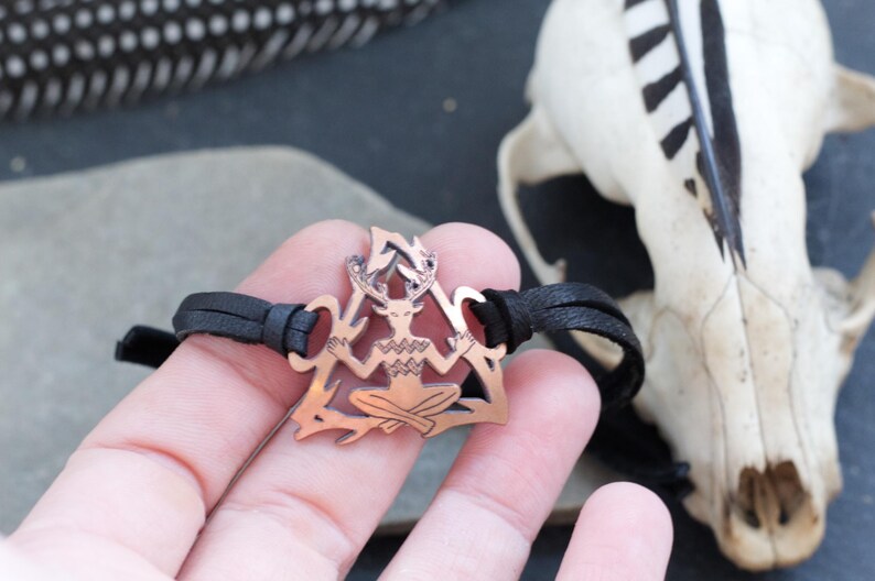 May include: A copper-colored bracelet with a leather cord and a pendant featuring a stylized figure with antlers, sitting in a triangular shape.