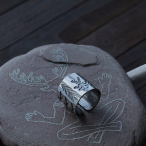 May include: A silver ring with a design of a deer and arrows. The ring is sitting on a stone with a carving of a deer.
