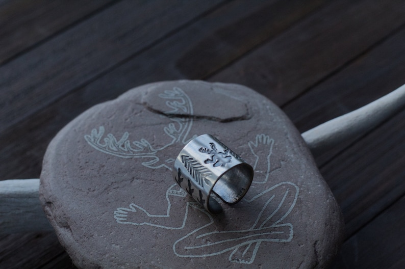 May include: A silver ring with a design of a deer and arrows. The ring is sitting on a stone with a carving of a deer.