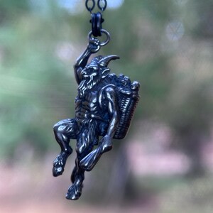May include: A silver-toned pendant necklace featuring a detailed depiction of a satyr carrying a basket. The satyr is posed in a climbing position, with a beard and horns. The pendant hangs from a dark chain.
