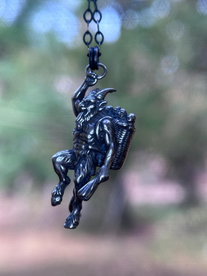 May include: A silver-toned pendant necklace featuring a detailed depiction of a satyr carrying a basket. The satyr is posed in a climbing position, with a beard and horns. The pendant hangs from a dark chain.