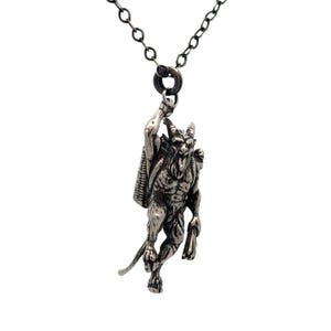 May include: A silver-toned pendant necklace featuring a detailed depiction of a horned, demonic figure with a satchel. The pendant is suspended from a dark chain, creating a striking contrast against the white background. The necklace is a gothic-themed accessory.