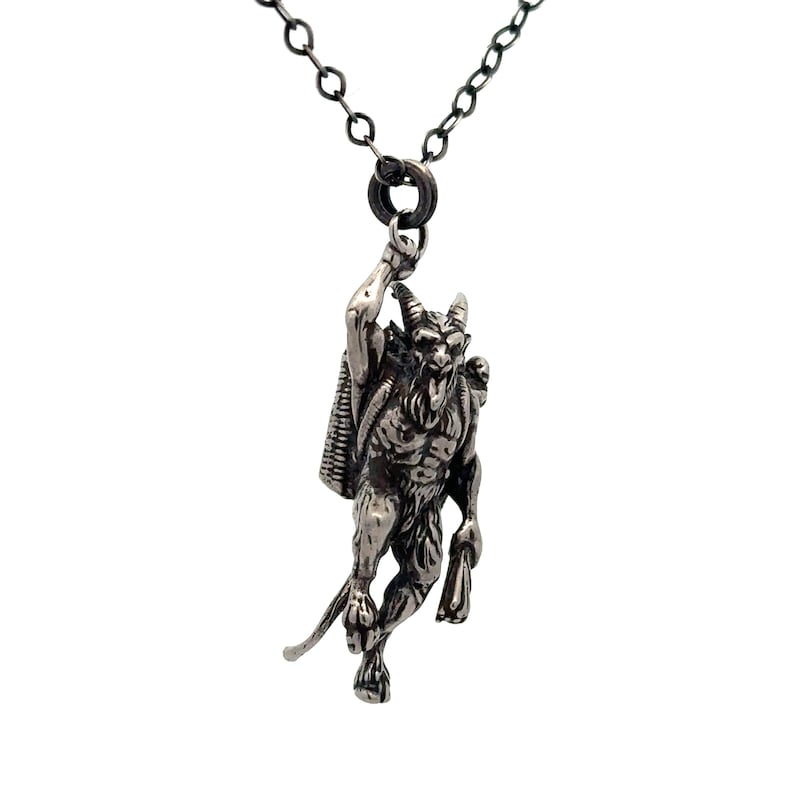 May include: A silver-toned pendant necklace featuring a detailed depiction of a horned, demonic figure with a satchel. The pendant is suspended from a dark chain, creating a striking contrast against the white background. The necklace is a gothic-themed accessory.
