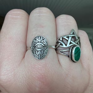 May include: Two silver rings are displayed on a hand. One ring features an oval face with a detailed, stylized design. The other ring has a Celtic knot design and a green oval stone. The rings are worn on different fingers.