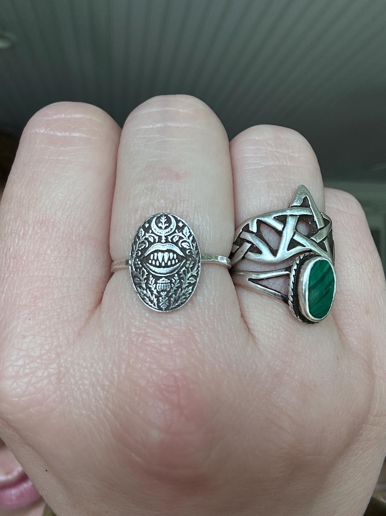 May include: Two silver rings are displayed on a hand. One ring features an oval face with a detailed, stylized design. The other ring has a Celtic knot design and a green oval stone. The rings are worn on different fingers.