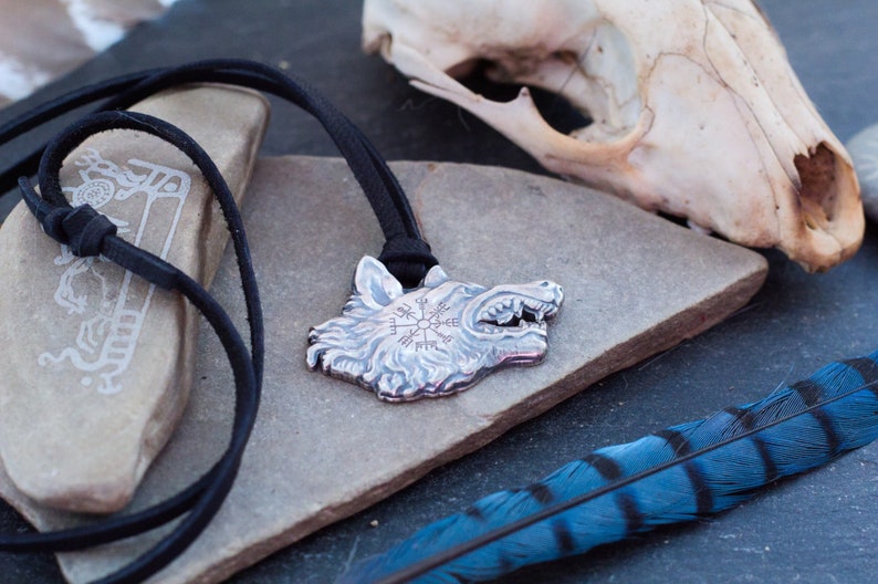 May include: A silver wolf head pendant with a compass design on the forehead, hanging on a black leather cord. The pendant is resting on a gray stone with a blue and black feather in the background.