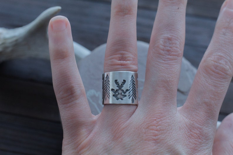 May include: A silver ring with a design of a deer with antlers and pine trees on either side.