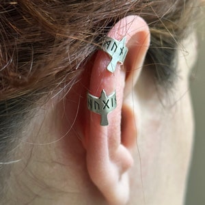 May include: Two silver ear cuffs with a bird design. The cuffs are inscribed with the text "NNXIX" and "ANXIX".