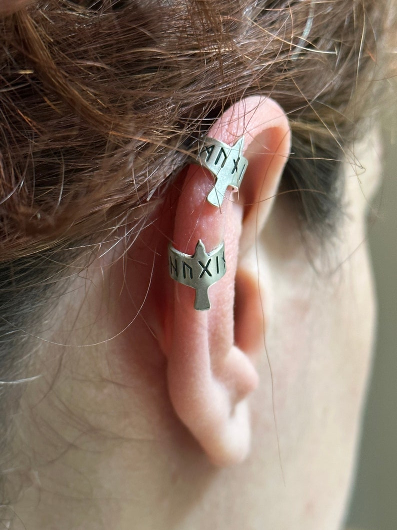 May include: Two silver ear cuffs with a bird design. The cuffs are inscribed with the text "NNXIX" and "ANXIX".