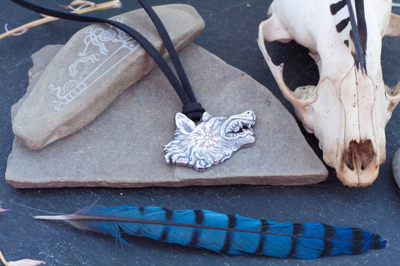 May include: Silver wolf head pendant with a compass design on the forehead, hanging from a black cord on a gray stone surface. A blue and black feather and a white animal skull are also in the image.