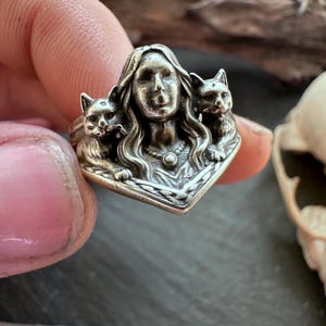 K&ouml;nnte beinhalten: Ein silberner Ring mit dem Gesicht einer Frau, flankiert von zwei Katzen. Der Ring weist aufwendige Details auf, darunter das Haar der Frau und das Fell der Katzen. Ein einzigartiges Schmuckst&uuml;ck.
