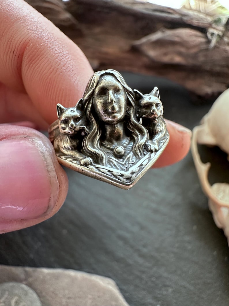 K&ouml;nnte beinhalten: Ein silberner Ring mit dem Gesicht einer Frau, flankiert von zwei Katzen. Der Ring weist aufwendige Details auf, darunter das Haar der Frau und das Fell der Katzen. Ein einzigartiges Schmuckst&uuml;ck.