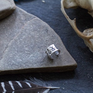 May include: A silver ring with a design of trees and leaves on the band. The ring is on a gray stone surface.
