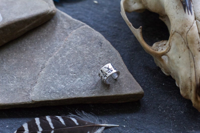 May include: A silver ring with a design of trees and leaves on the band. The ring is on a gray stone surface.