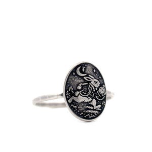 May include: A silver ring featuring an oval pendant with a detailed engraving of a rabbit wearing a waistcoat and holding a pocket watch, set against a starry night sky. The ring has a textured band.