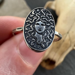 May include: A silver ring featuring an oval medallion with a detailed depiction of Medusa's face, surrounded by snakes. The ring has a textured band and a dark, antique finish, showcasing the intricate design.