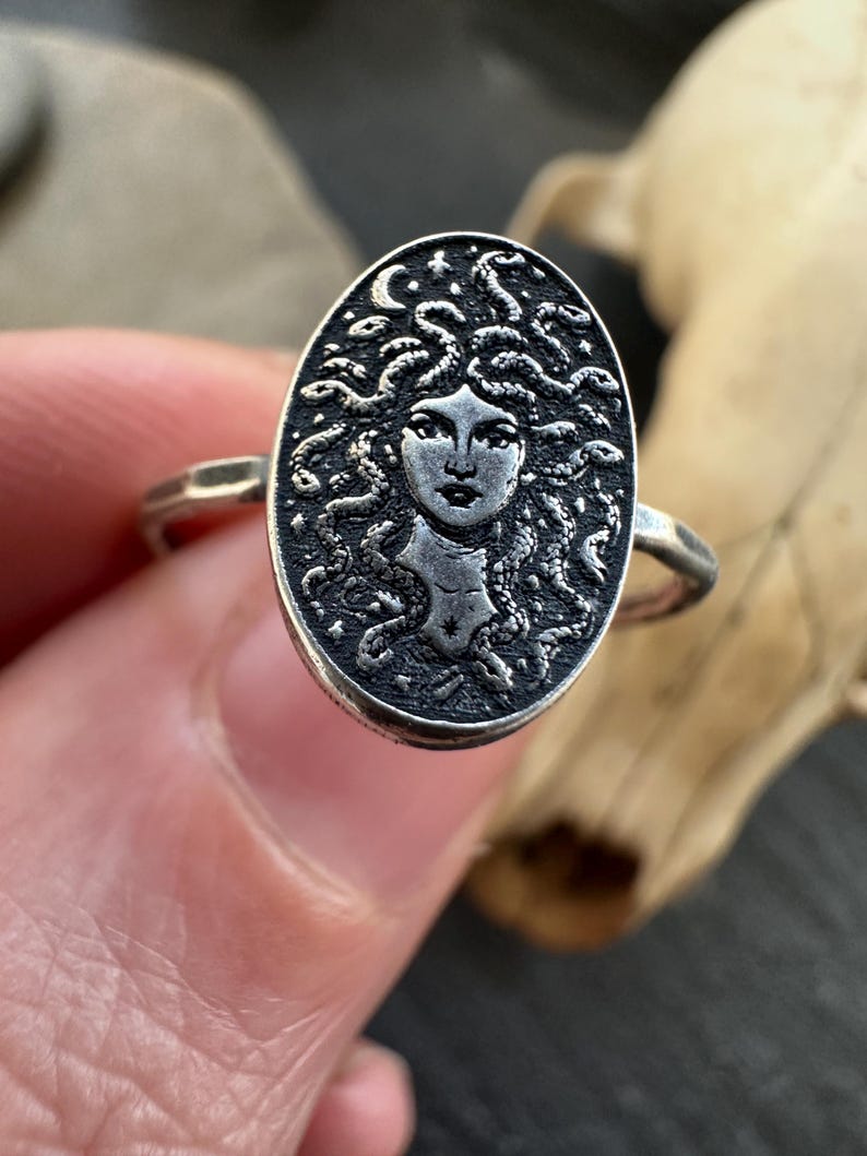 May include: A silver ring featuring an oval medallion with a detailed depiction of Medusa's face, surrounded by snakes. The ring has a textured band and a dark, antique finish, showcasing the intricate design.