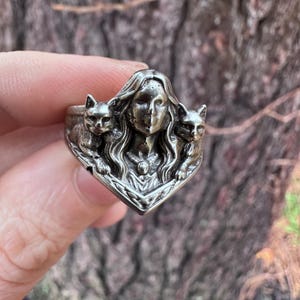 K&ouml;nnte beinhalten: Ein silberner Ring mit dem Gesicht einer Frau, flankiert von zwei Katzenfiguren. Der Ring hat aufwendige Details und einen Vintage-Look. Ein Schmuckst&uuml;ck f&uuml;r Katzenliebhaber und alle, die einzigartige Designs sch&auml;tzen. Das Design des Rings ist aufwendig.