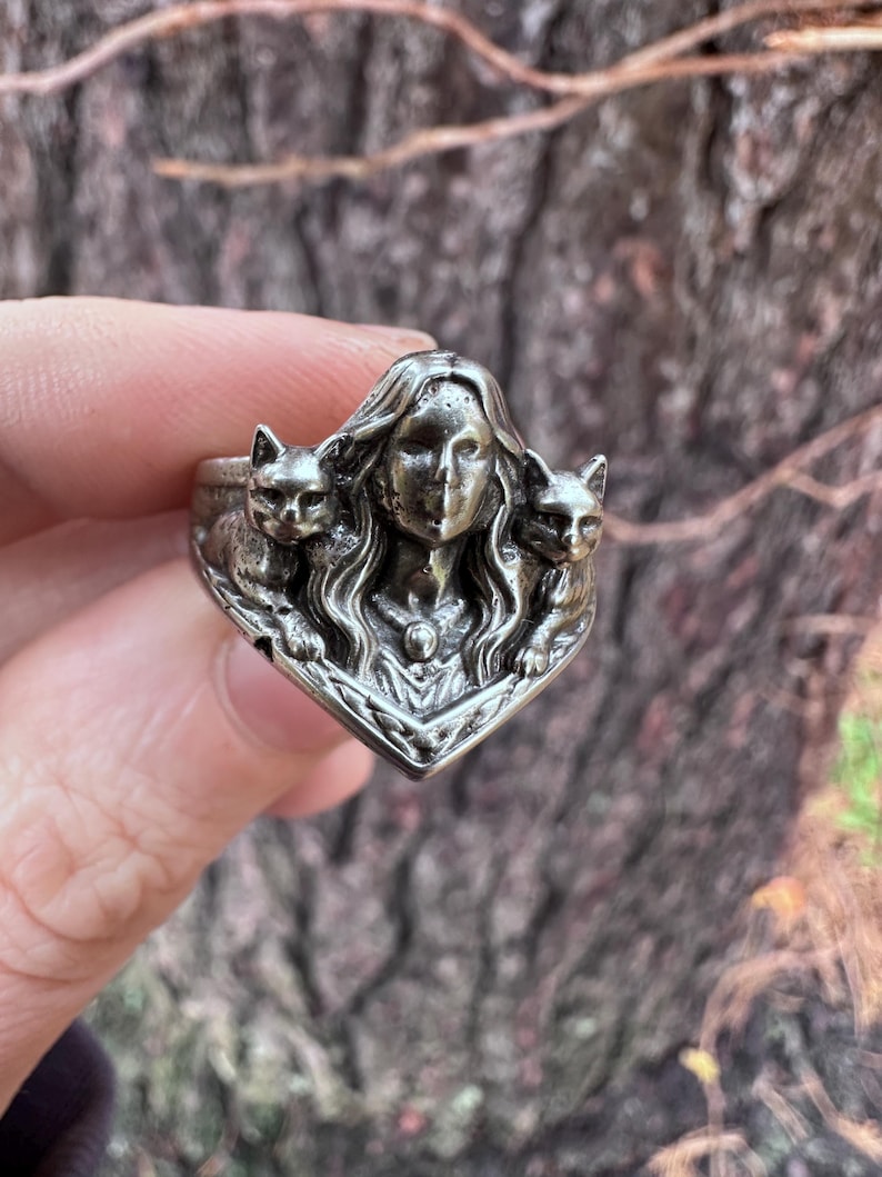 K&ouml;nnte beinhalten: Ein silberner Ring mit dem Gesicht einer Frau, flankiert von zwei Katzenfiguren. Der Ring hat aufwendige Details und einen Vintage-Look. Ein Schmuckst&uuml;ck f&uuml;r Katzenliebhaber und alle, die einzigartige Designs sch&auml;tzen. Das Design des Rings ist aufwendig.