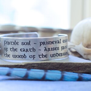 May include: A silver metal bracelet with an engraved quote: "Fertile soil - primeval essence of the earth - arises from the womb of the underworld."