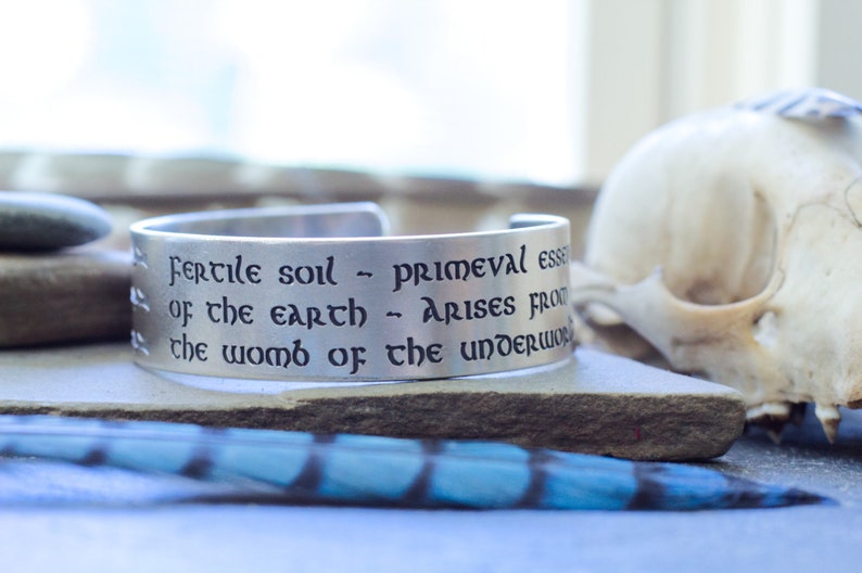 May include: A silver metal bracelet with an engraved quote: "Fertile soil - primeval essence of the earth - arises from the womb of the underworld."
