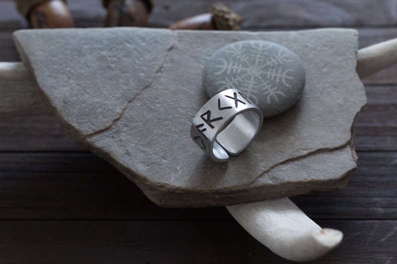 May include: A silver ring with a band of runes inscribed on it. The ring is sitting on a gray stone with a carved symbol.