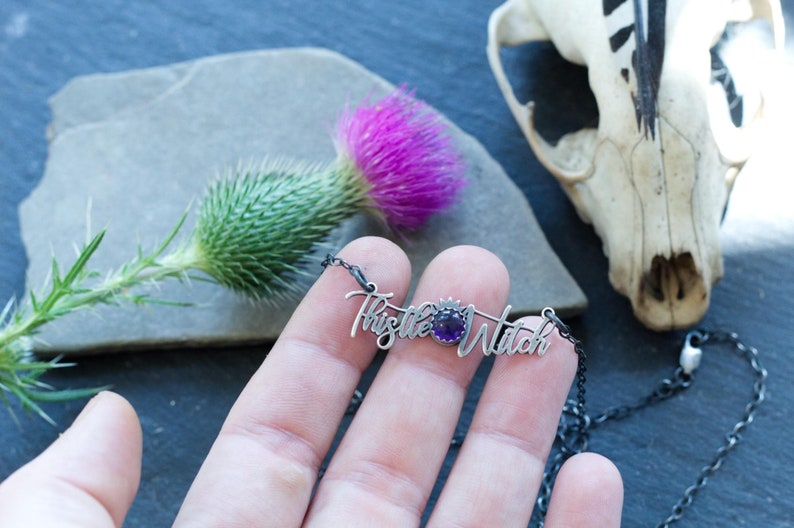 May include: A silver necklace with a purple gemstone and the words "Thistle Witch" written in cursive.