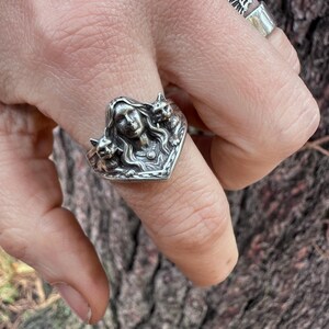 K&ouml;nnte beinhalten: Ein silberner Ring mit dem Gesicht einer Frau und zwei Katzen. Der Ring hat detaillierte Merkmale und ein herzf&ouml;rmiges Design. Ein weiterer Ring mit einem Fl&uuml;geldesign ist ebenfalls sichtbar.