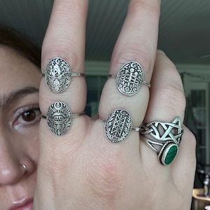 May include: A hand displaying five silver rings. Four rings have oval faces with detailed designs, including a crescent moon, skulls, and geometric patterns. The fifth ring features a green malachite stone and a Celtic knot design.
