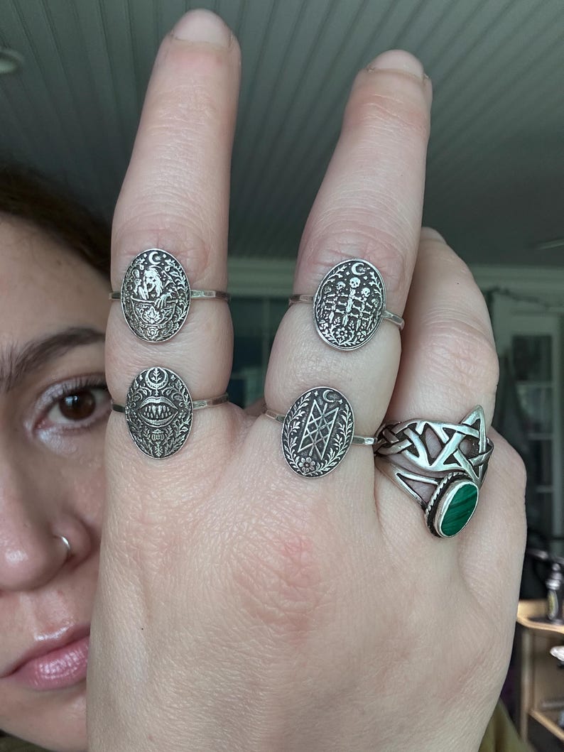 May include: A hand displaying five silver rings. Four rings have oval faces with detailed designs, including a crescent moon, skulls, and geometric patterns. The fifth ring features a green malachite stone and a Celtic knot design.