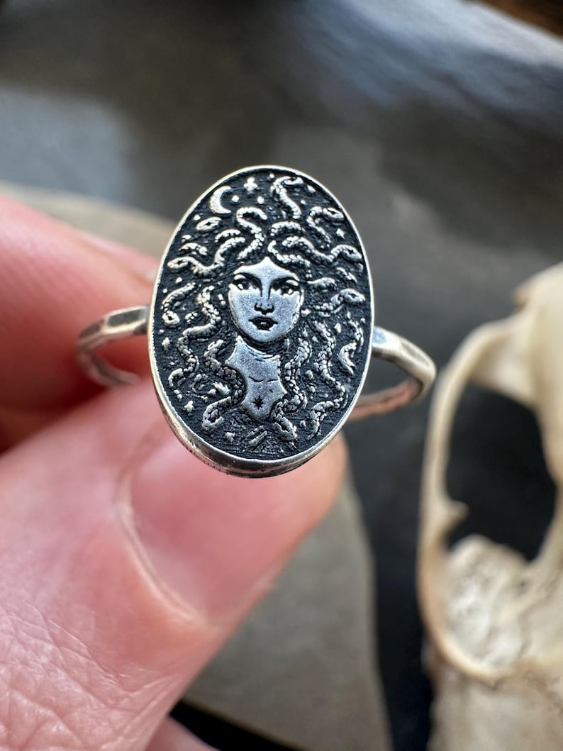 May include: A silver ring featuring an oval medallion with a detailed depiction of Medusa's face, complete with coiling snakes for hair. The ring has a textured band and a dark, antique finish, showcasing the intricate design.