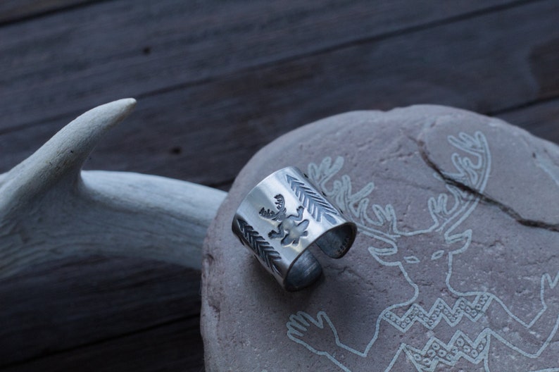 May include: A silver ring with a deer design on a stone background with a deer carving.