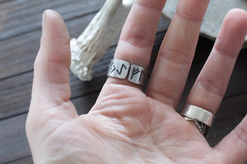May include: A silver ring with a band that is engraved with runes. The ring is worn on the middle finger of a hand.