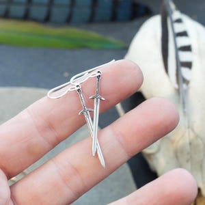 Peut inclure: Une paire de boucles d'oreilles en argent en forme d'&eacute;p&eacute;e. Les &eacute;p&eacute;es sont d&eacute;taill&eacute;es et ont une pointe ac&eacute;r&eacute;e. Les boucles d'oreilles pendent de la main d'une personne.