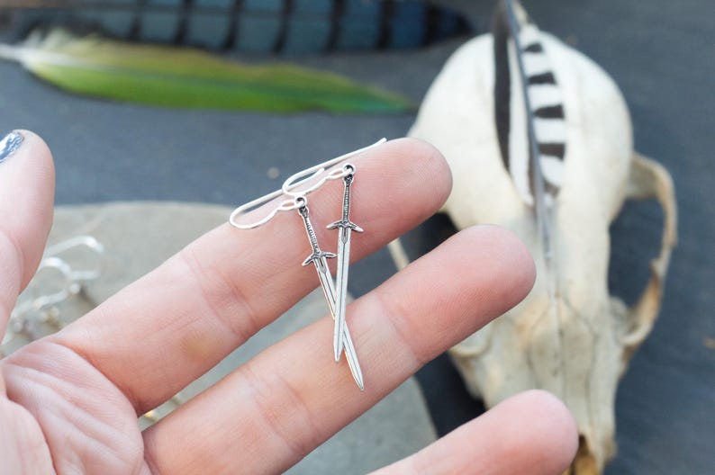 Peut inclure: Une paire de boucles d'oreilles en argent en forme d'&eacute;p&eacute;e. Les &eacute;p&eacute;es sont d&eacute;taill&eacute;es et ont une pointe ac&eacute;r&eacute;e. Les boucles d'oreilles pendent de la main d'une personne.