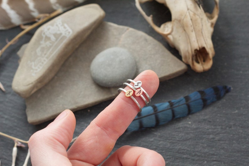 May include: Three silver rings with different rune symbols on the top of each ring. The rings are stacked on a finger.