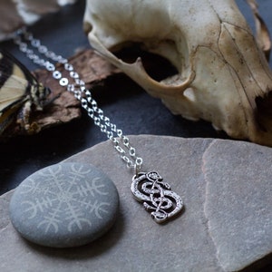 May include: Silver chain necklace with a silver pendant featuring a knot design. The necklace is displayed on a gray stone with a carved symbol and a white animal skull.