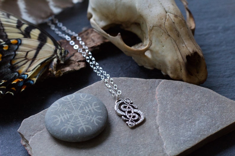 May include: Silver chain necklace with a silver pendant featuring a knot design. The necklace is displayed on a gray stone with a carved symbol and a white animal skull.