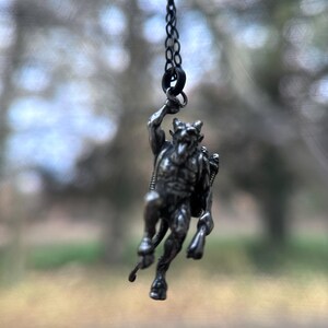 May include: A silver-toned pendant necklace featuring a detailed depiction of a horned, humanoid figure with a tail. The pendant is suspended from a delicate chain, showcasing the intricate design of the mythological creature.
